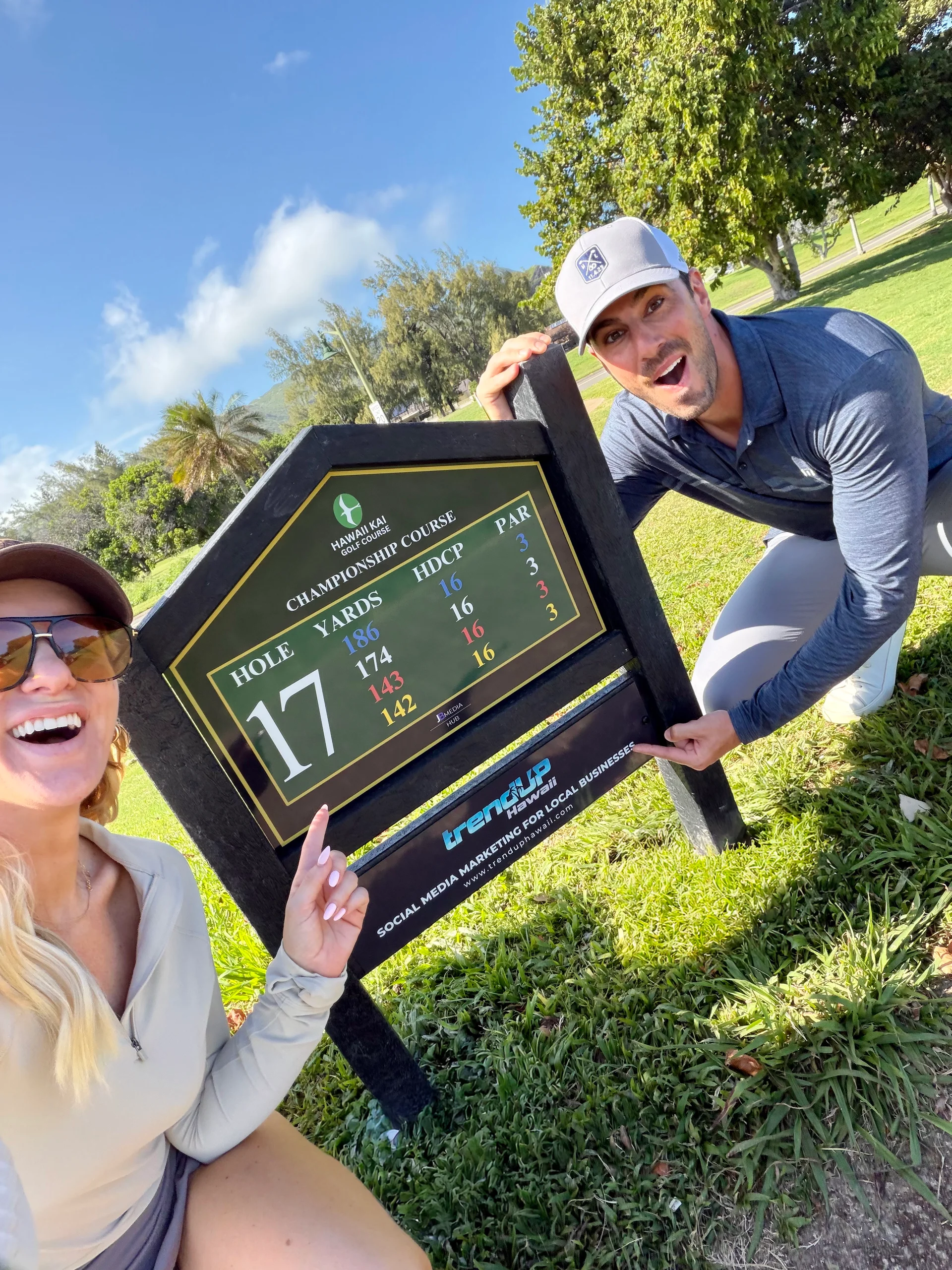 Two people smiling beside the Hawaii Kai Golf Course tee sign and TrendUP Hawaii sponsor panel