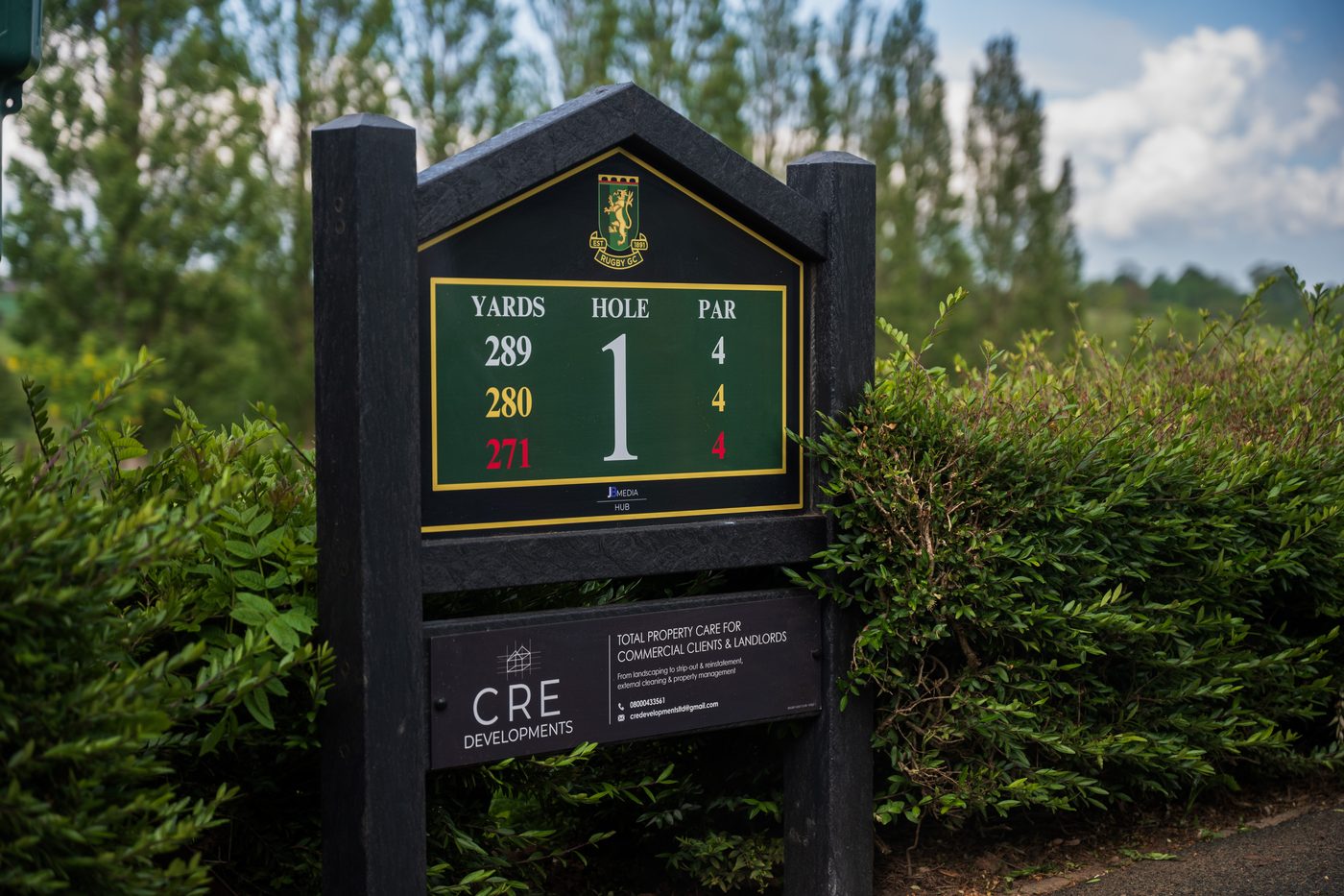 A JB Golf Group tee box sign on a golf course, with a local business's sponsor panel beneath it